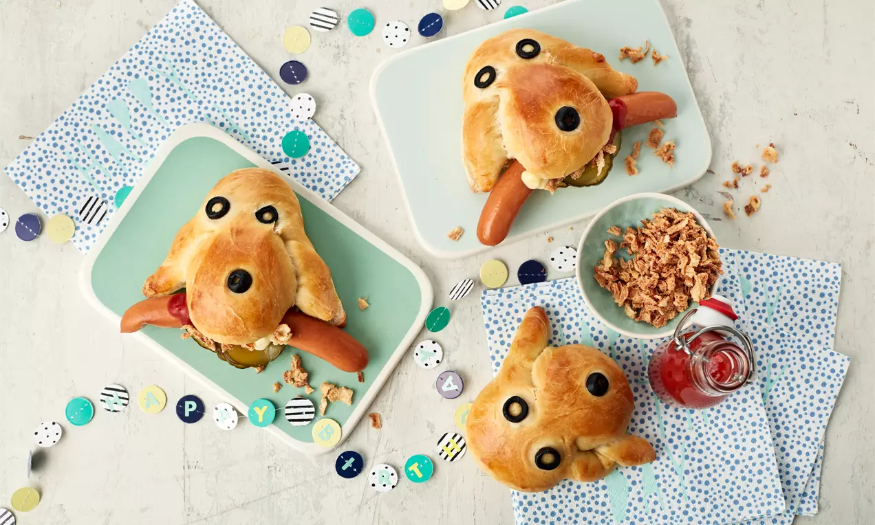Goldbraune Hot-Dog-Brötchen in Hundeform mit schwarzen Olivenaugen und Würstchenfüllung auf pastellfarbenen Tellern, dekoriert mit bunten Papierkonfetti