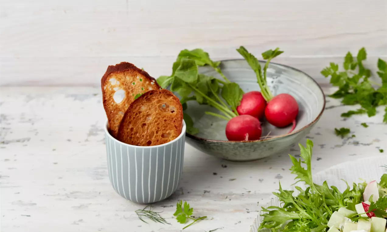 Brotchips in einem Streifenbecher und einem rustikalen Schälchen arrangiert, daneben Radieschen und angeschnitten Salatteller
