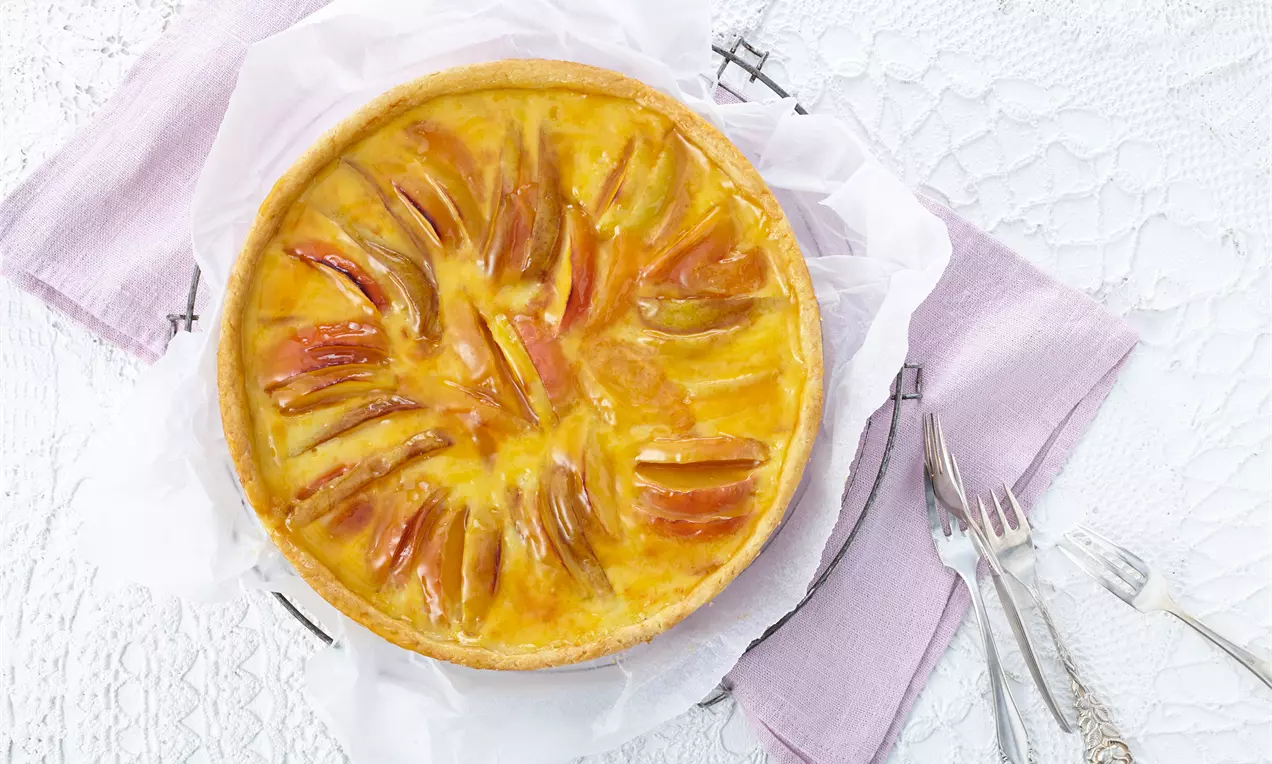 Französischer Obstkuchen mit goldgelber Glasur und dekorativ angeordneten Apfelscheiben auf weißem Teller mit lilafarbenen Servietten