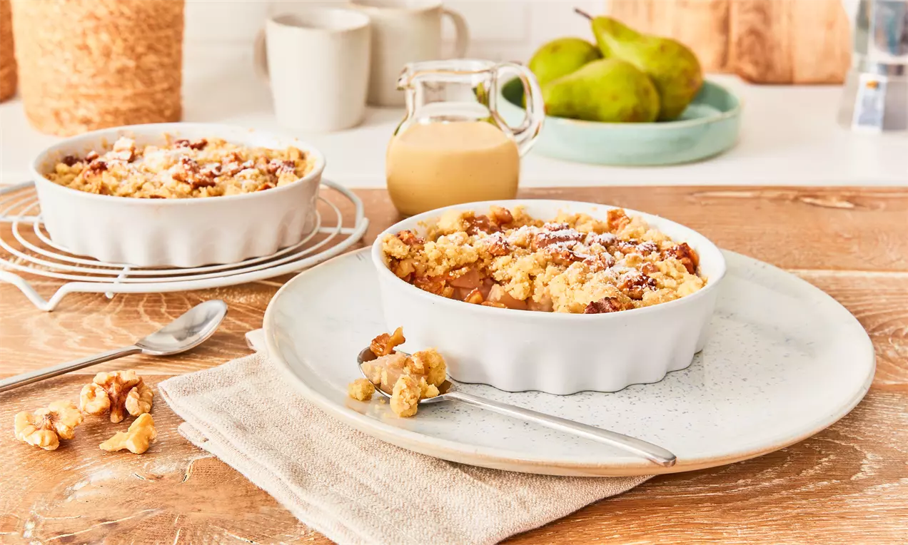 Veganer Birnen-Crumble mit goldbrauner Kruste und Streuseln in einer weißen Auflaufform, garniert mit Walnüssen auf einem Holztisch