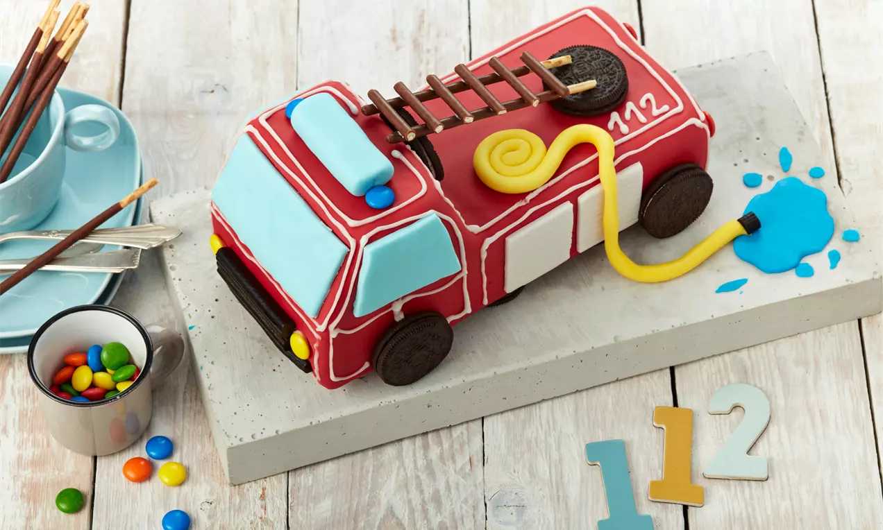 Feuerwehrauto-Kuchen mit roter Glasur und Zuckerdekoration, gelbem Schlauch, Eistütenleiter und blauer Wasserpfütze auf hellem Untergrund