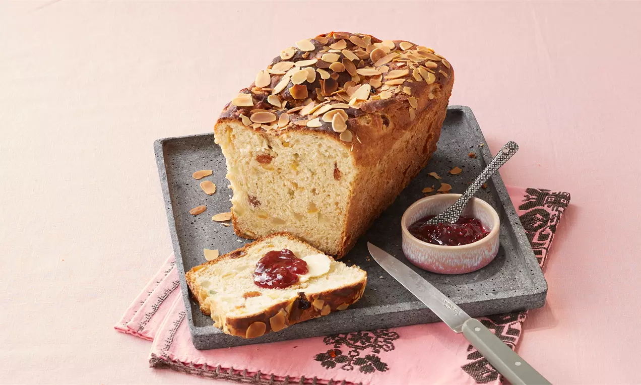 Norwegisches Weihnachtsbrot mit goldbrauner Kruste und Mandelblättchen, angeschnitten auf grauer Platte, daneben Marmelade und Messer
