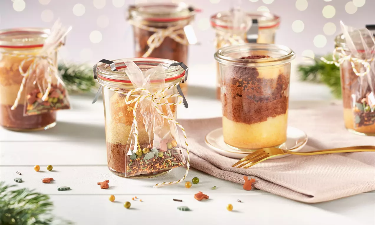 Schichtkuchen im Glas mit dunkler und heller Teigschicht, dekoriert mit Schleifen, umgeben von Tannenzweigen auf hellem Tisch