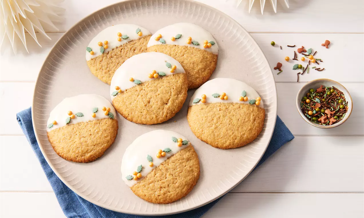 Runde Lebkuchen-Cookies mit weißem Zuckerguss und bunter Streudekoration auf einem hellen Teller, arrangiert auf blauem Stoff