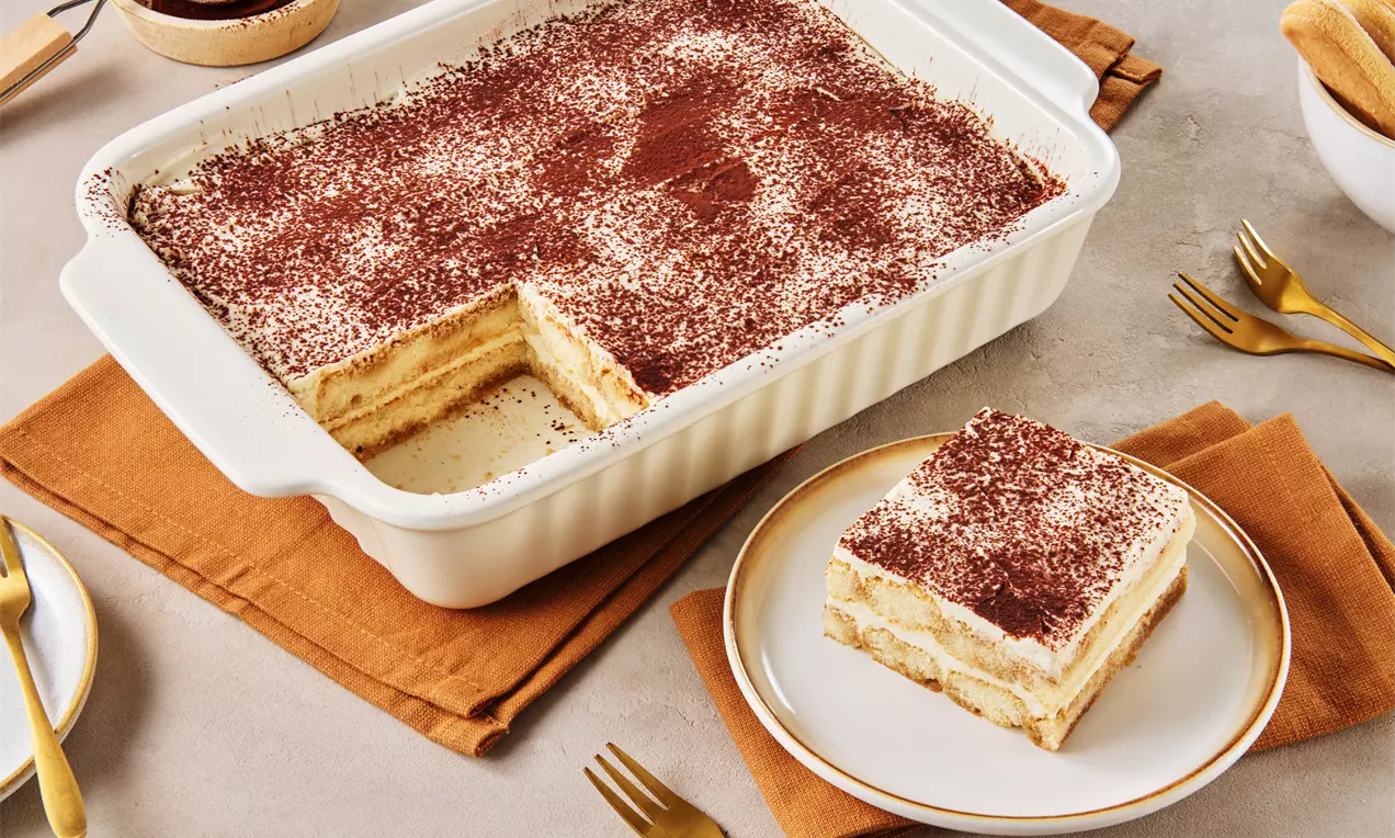 Klassisches Tiramisu in einer weißen Auflaufform mit Kakaoschicht, angeschnitten und ein Stück auf einem Teller mit goldener Gabel angerichtet