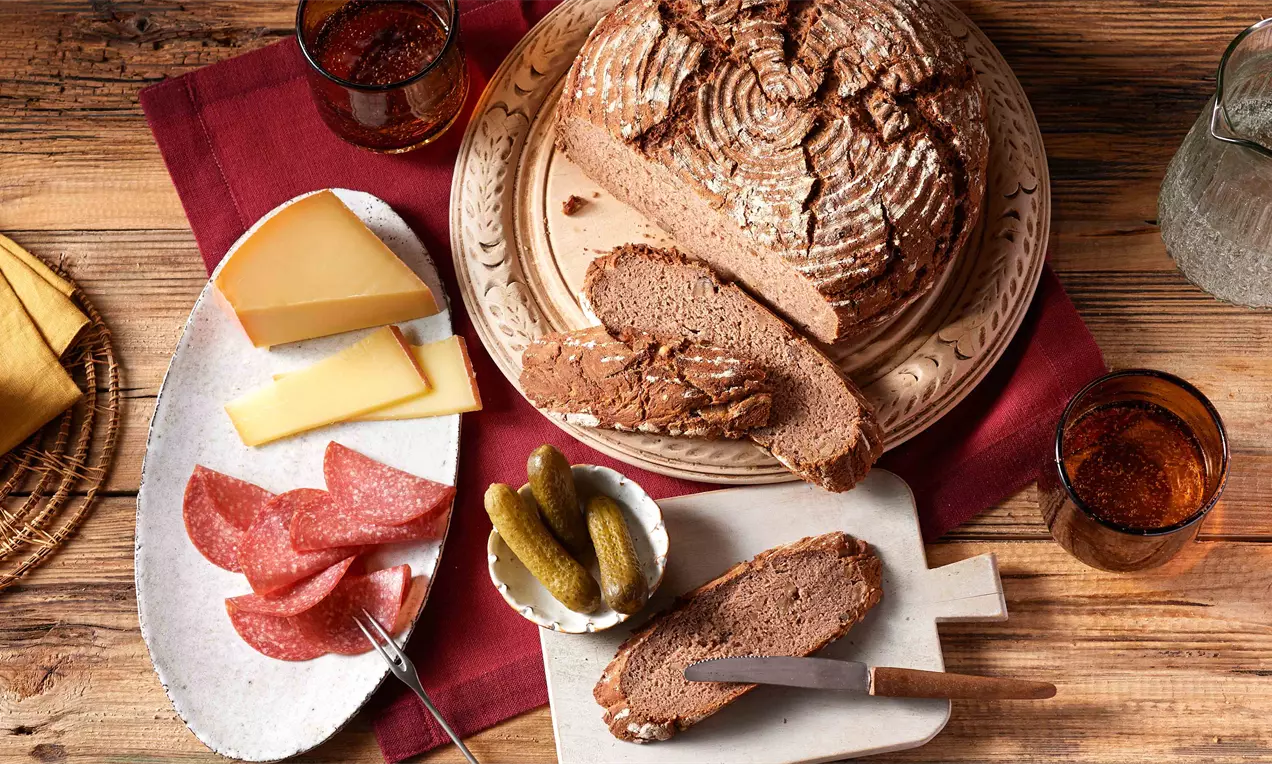 Rustikales Kastanienbrot mit rissiger Kruste, angeschnitten auf einem Holztisch neben Wurst, Käse, Cornichons und Glasgeschirr