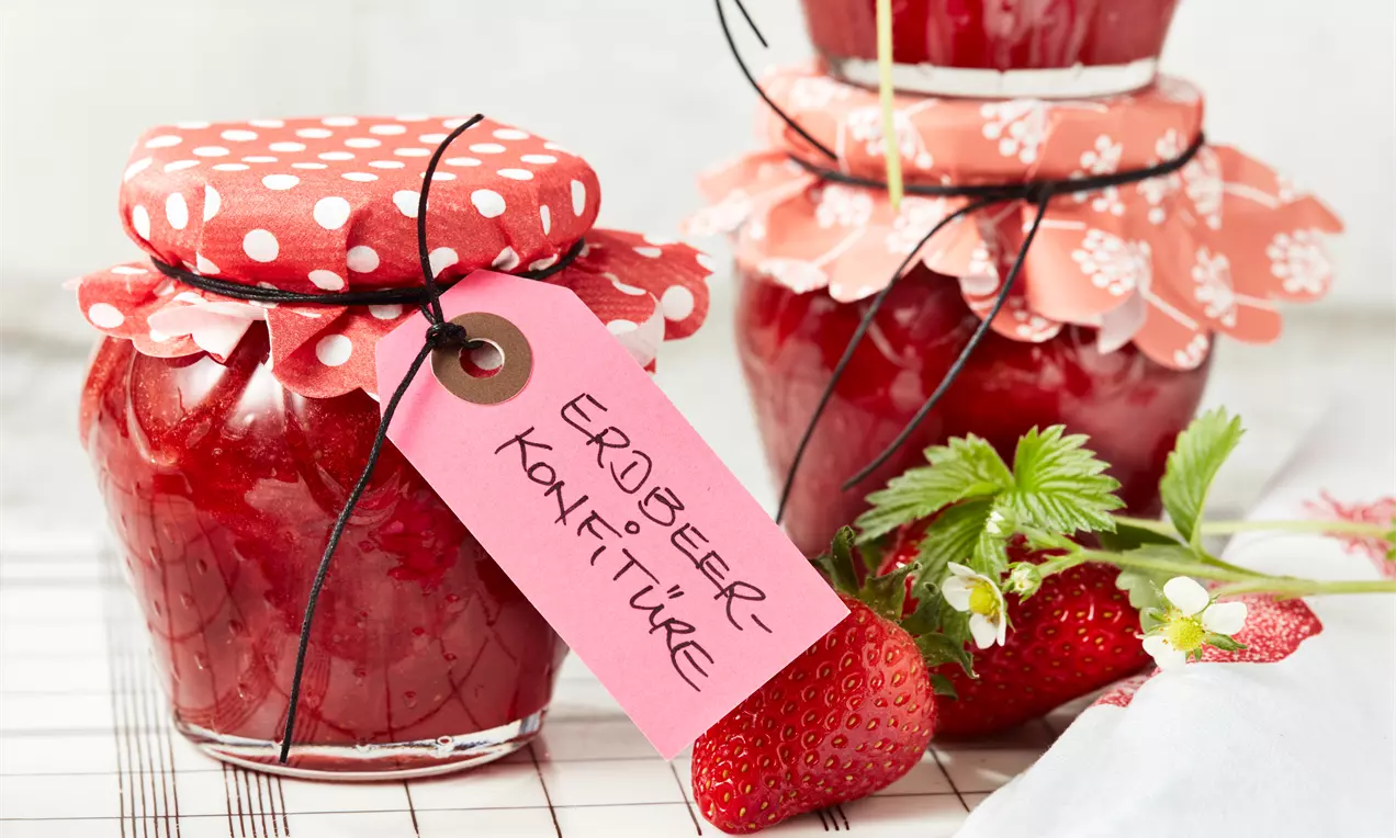 Gläser mit leuchtend roter Erdbeer-Konfitüre, abgedeckt mit gemusterten Stoffdeckeln, neben frischen Erdbeeren und grünem Blattwerk