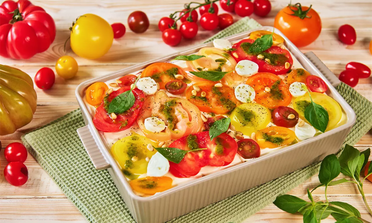 Schichten aus leuchtenden Tomatenscheiben, Mozzarella-Kugeln und Basilikumblättern, dekoriert mit grüner Sauce in einer rechteckigen Form