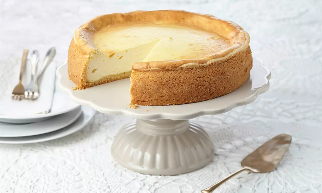 Goldbrauner Käsekuchen mit glatter, hellgelber Füllung auf weißer Tortenplatte, angerichtet neben Besteck und Teller auf Spitzenunterlage