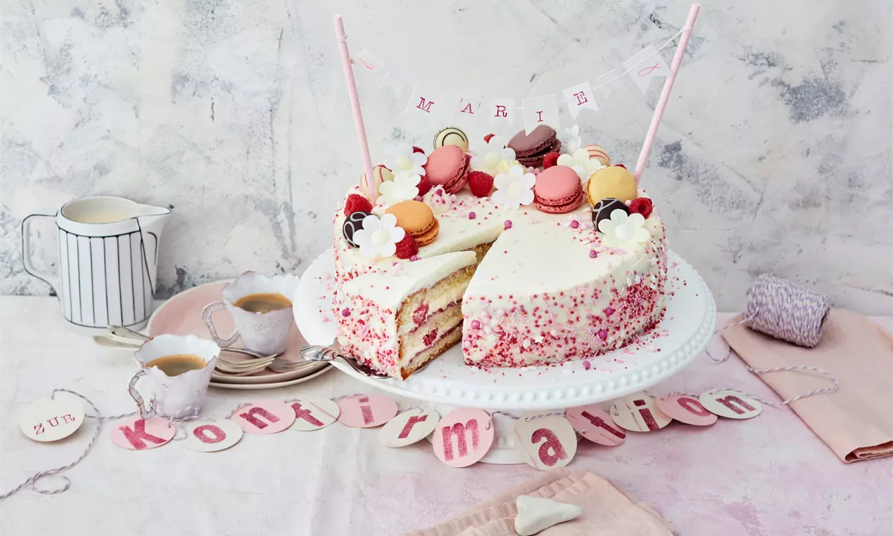 Runde weiße Torte mit rosa Streuseln, frischen Beeren, bunten Macarons und Zuckerdeko, auf einer Tortenplatte mit zarter Tischdekoration