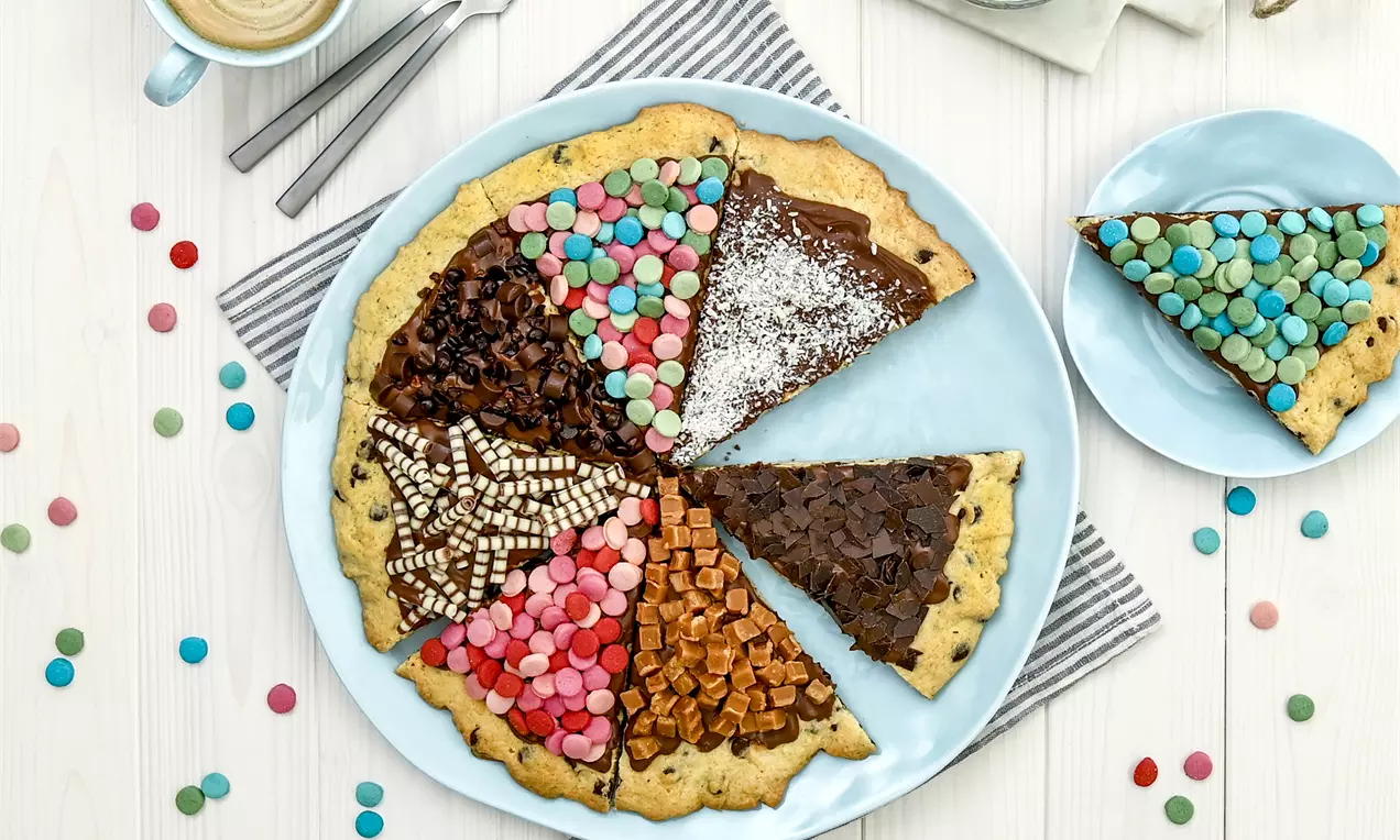 Runde Cookie-Pizza mit braunem Boden, in bunte Süßigkeiten-Segmente geteilt, auf weißem Teller mit gelber Serviette angerichtet