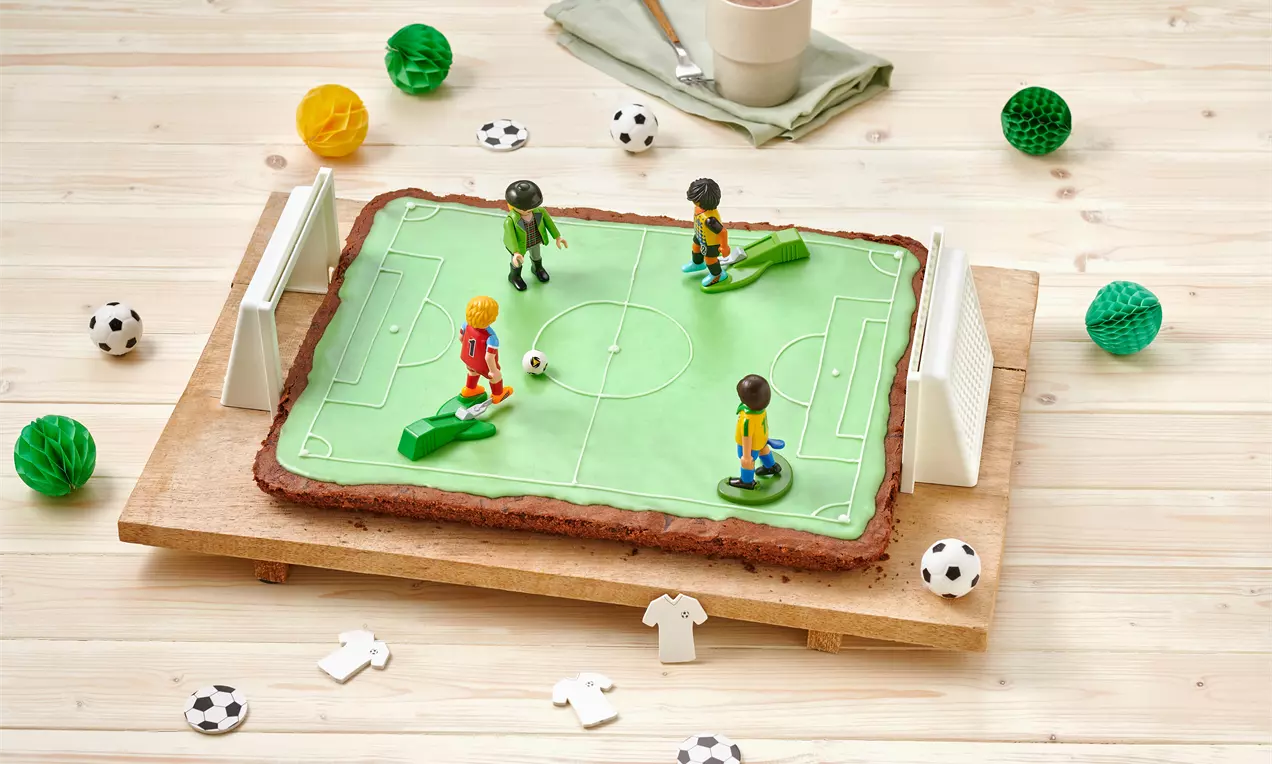Rechteckiger Fußballkuchen mit grüner Spielfelddecke, weißen Toren und Spielfiguren auf Holzbrett, umgeben von Deko-Fußbällen