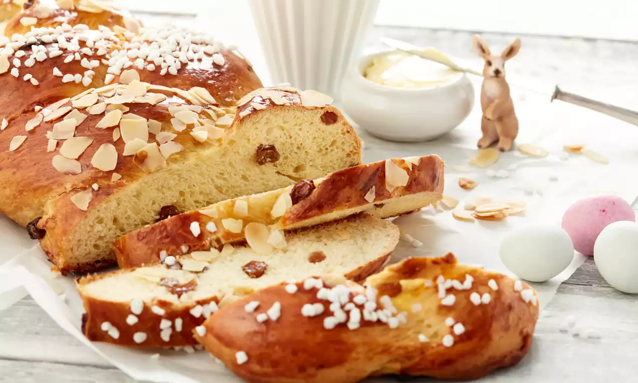 Goldbrauner Osterzopf mit Zuckerperlen und Mandelblättchen, locker geschnitten, daneben Dekoration mit Ostereiern und Hasenfigur auf hellem Holz