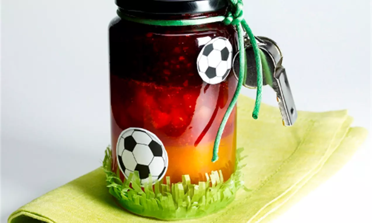 Glas mit dunkelroter Konfitüre und orangefarbenem Farbverlauf, verziert mit Fußballstickern und grünem Band auf grünem Stoff drapiert