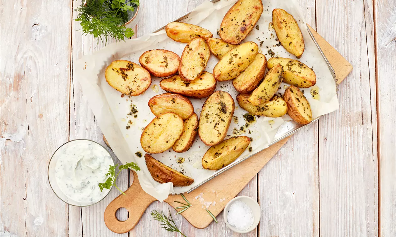 Goldbraune Ofenkartoffelhälften mit Kräutern bestreut auf Backpapier, daneben eine Schale mit weißem Kräuterquark und frischem Dill