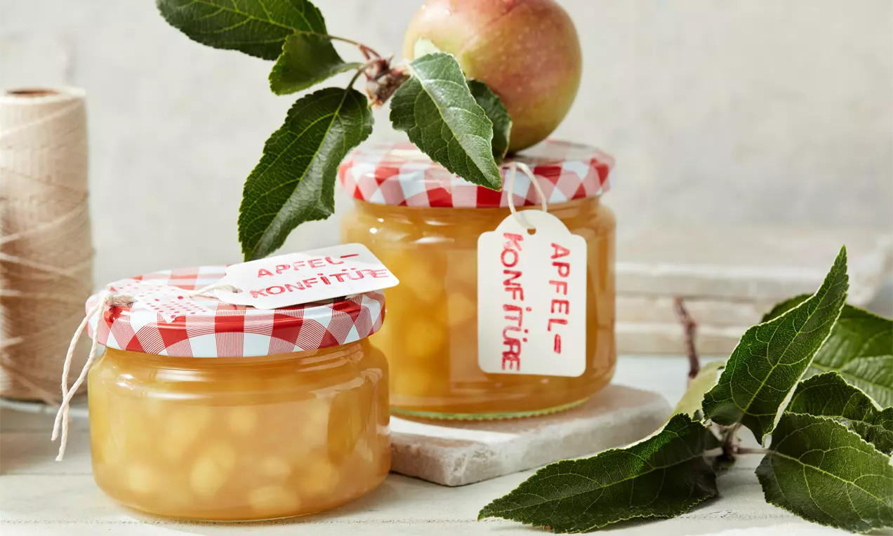 Apfelmarmelade in einem Glas und auf knusprigem Vollkornbrot, glänzende goldrote Konfitüre mit Apfelscheiben, Zimtstange und frischem Thymian