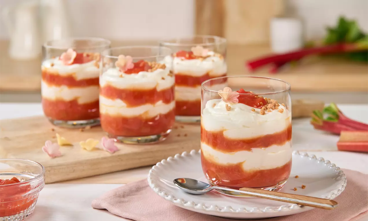 Schichtdessert aus weißer Creme und rot-orange Rhabarberkompott in einem Glas, garniert mit Streuseln und Blüten auf weißem Teller