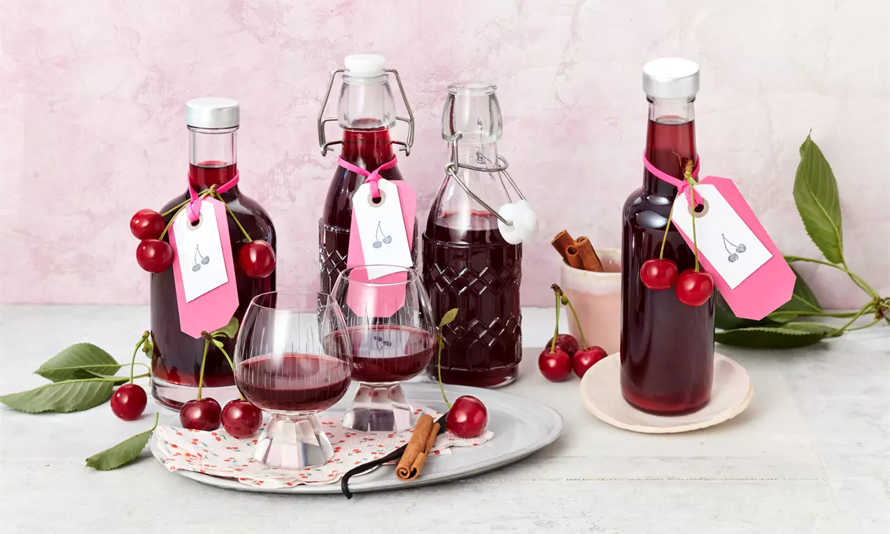Flaschen mit tiefrotem Kirschlikör und pinken Anhängern, umgeben von frischen Kirschen und Gläsern auf Tellern mit Zimtstangen