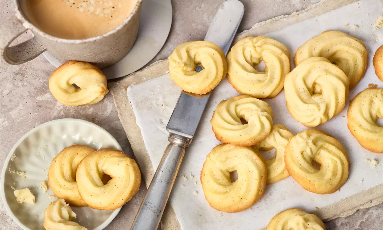 Butter Cookies auf einem weißen Teller: goldbraun, teils mit Puderzucker bestäubt, teils mit Schokotropfen, dekorativ gestapelt