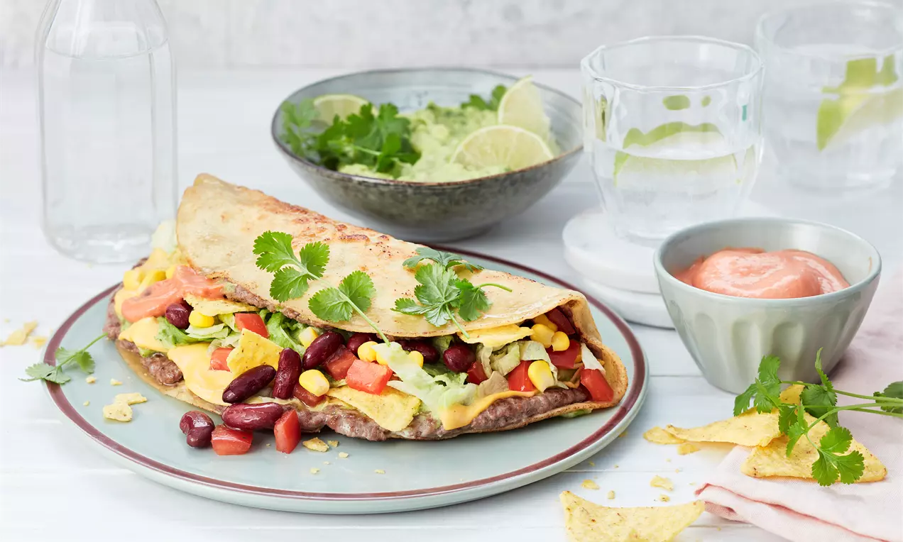 Ein Taco Burger aus goldbraunem Teig, gefüllt mit rotem Kidneybohnenmix, gelbem Mais, Avocadocreme und Koriander auf grauem Teller drapiert