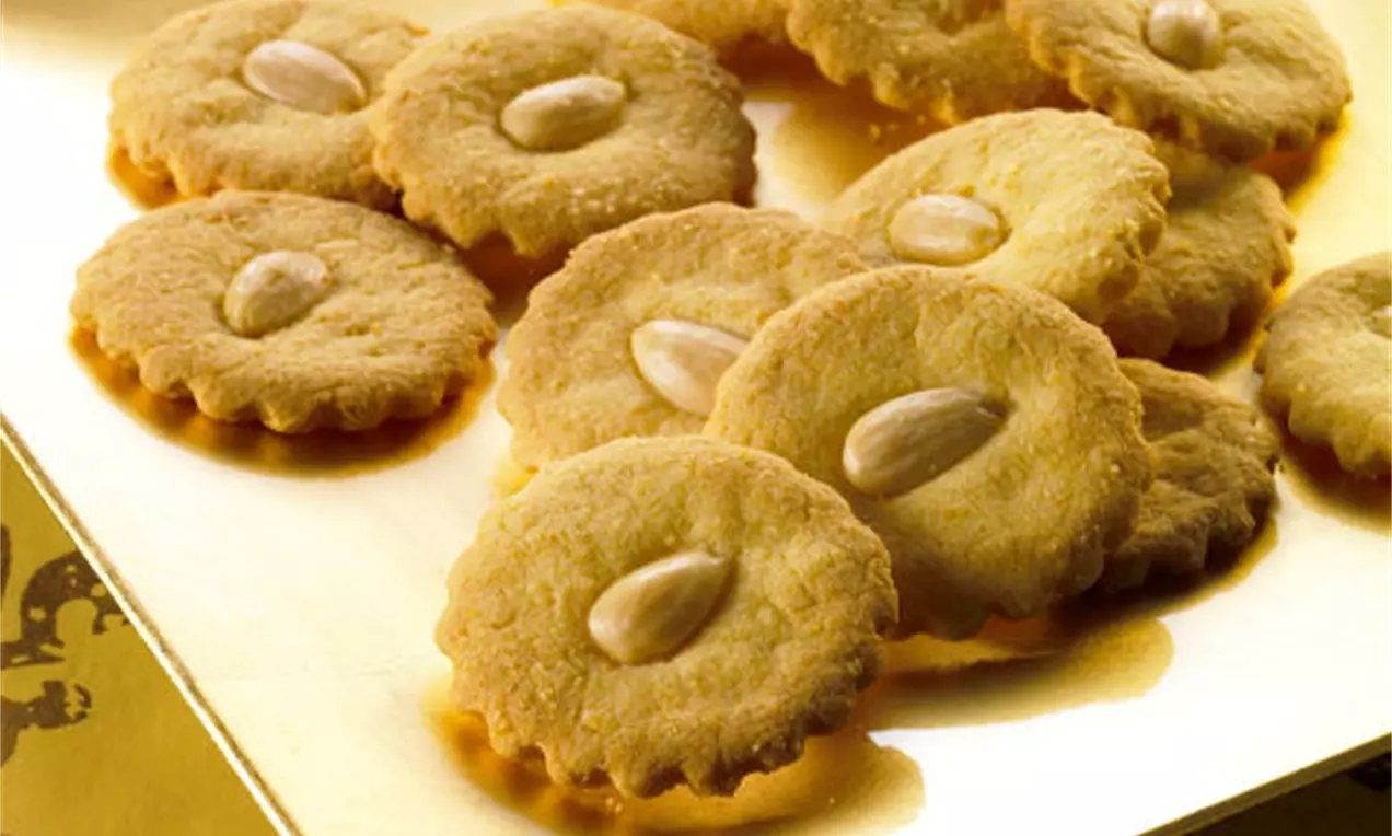 Goldfarbene, runde Plätzchen mit gezacktem Rand und glänzender Mandel in der Mitte auf einer schimmernden goldenen Platte arrangiert