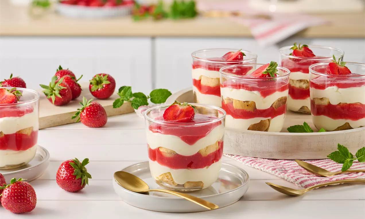 Gläser mit Schichten aus weißer Creme, roten Erdbeeren und Keksen, garniert mit Erdbeerstückchen und Minzblättern auf einem hellen Tisch