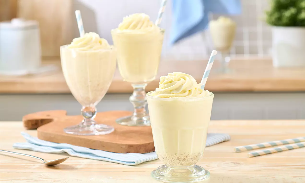 Drei Gläser mit cremigem Vanille-Milchshake, dekoriert mit Sahnehaube und Trinkhalm. Im Vordergrund ein Glas auf einem Holztisch, dahinter zwei Gläser auf einem Brett. Daneben gestreifte Strohhalme
