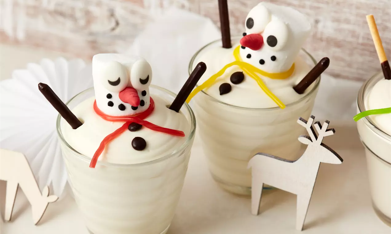 Winterliches Dessert mit weiß-cremiger Füllung in einem Glas, dekoriert mit Marshmallow-Schneemännern, bunten Bändern und Schokoladenelementen