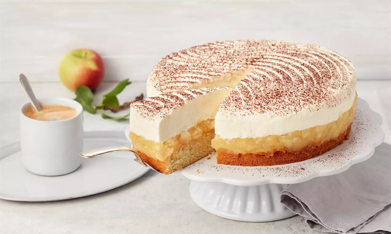 Apfeltorte mit hellem Boden, goldgelber Apfelschicht und weißer Cremeschicht, bestäubt mit Kakao, auf weißer Tortenplatte serviert
