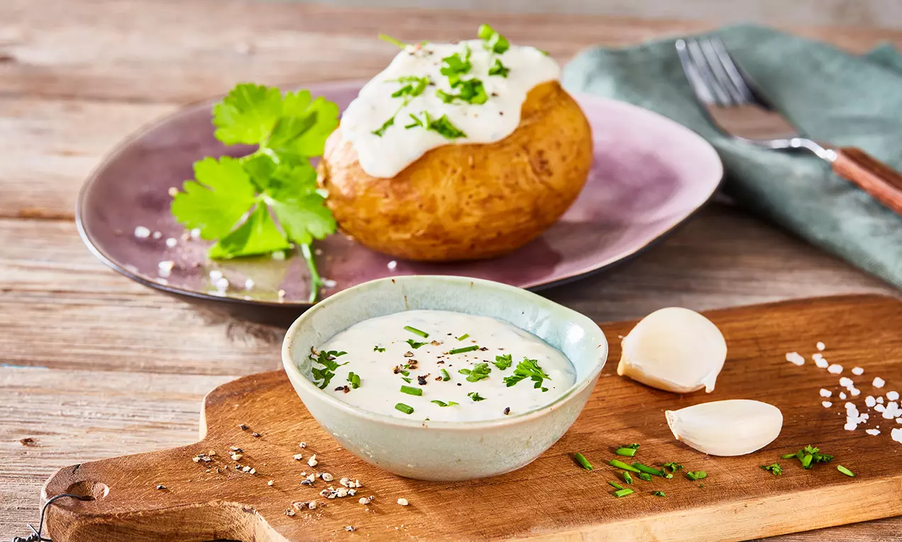 Eine goldbraune Ofenkartoffel mit weißer Sour-Cream und grünen Kräutern auf violettem Teller neben Schale mit Dip auf Holzbrett