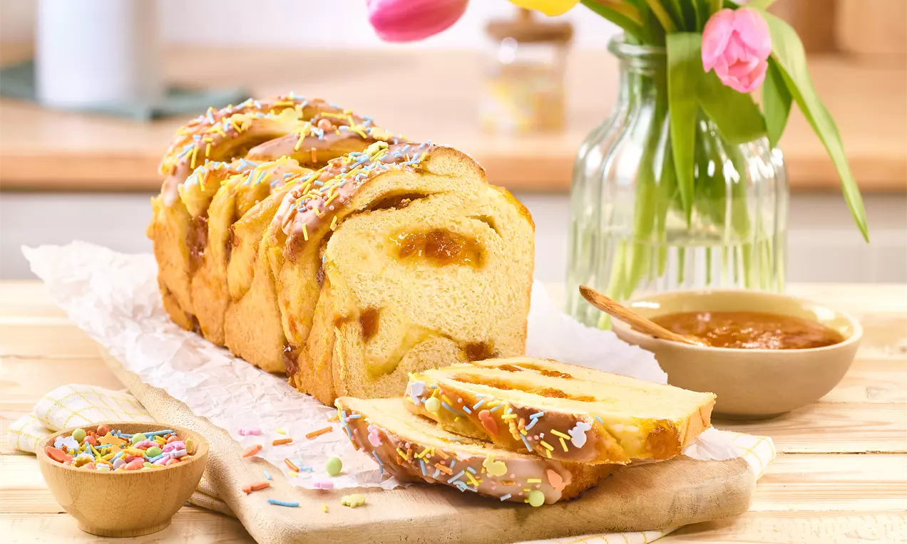 Geflochtene Babka mit goldbrauner Kruste und Marmeladenfüllung auf Holzbrett, dekoriert mit Zuckerstreuseln, daneben Tulpen und Marmeladenschale