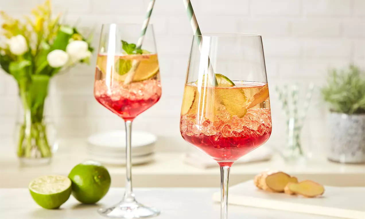 Zwei elegante Gläser mit roséfarbenem Getränk, Eiswürfeln, Limettenscheiben und Ingwerstücken, dekoriert mit Papierstrohhalmen