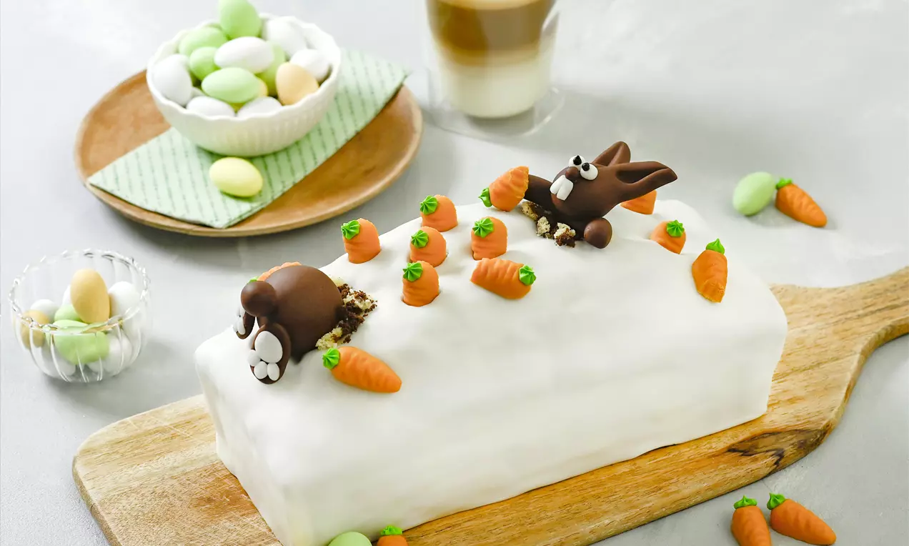 Rechteckiger Möhren-Marmorkuchen mit weißer Glasur, dekoriert mit kleinen Fondanthäschen und Möhren auf einem Holzbrett angerichtet
