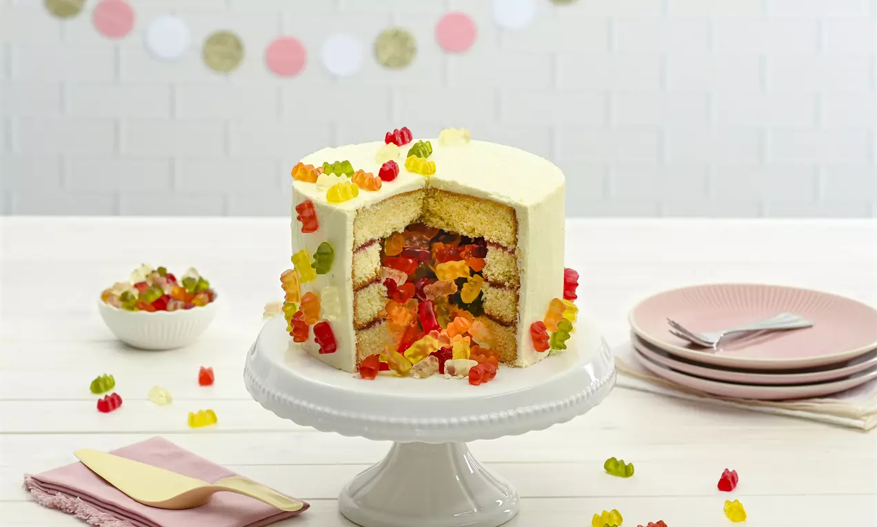Goldbären-Torte mit weißem Überzug, gefüllt mit bunten Gummibärchen, auf einer weißen Tortenplatte und Tisch mit rosa Geschirr drapiert