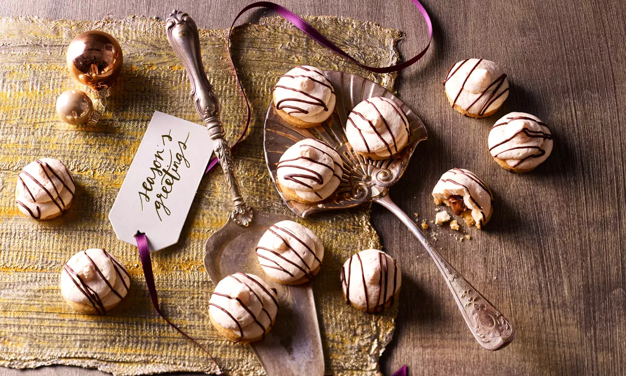 Nougatwölkchen mit heller, luftiger Oberfläche und dunkler Schokoladenverzierung auf goldener Tischdecke, umgeben von Besteck und Deko-Elementen
