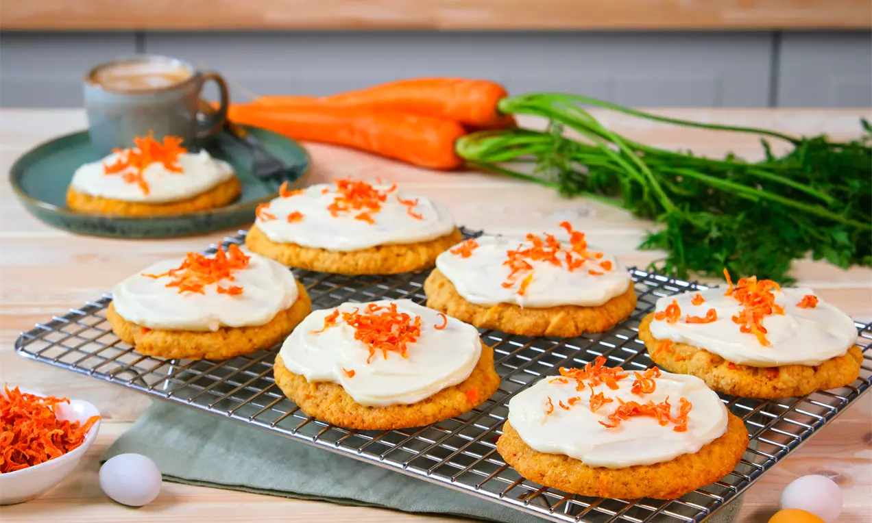 Runde Cookies mit cremefarbigem Frosting und geraspelten Karotten, auf einem Gitter serviert, daneben frische Karotten und eine Tasse Kaffee