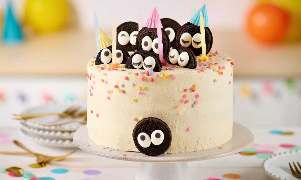 Kindergeburtstagstorte mit heller Creme, bunten Streuseln, schokoladigen Augen-Dekoren und Partyhütchen auf einer weißen Tortenplatte
