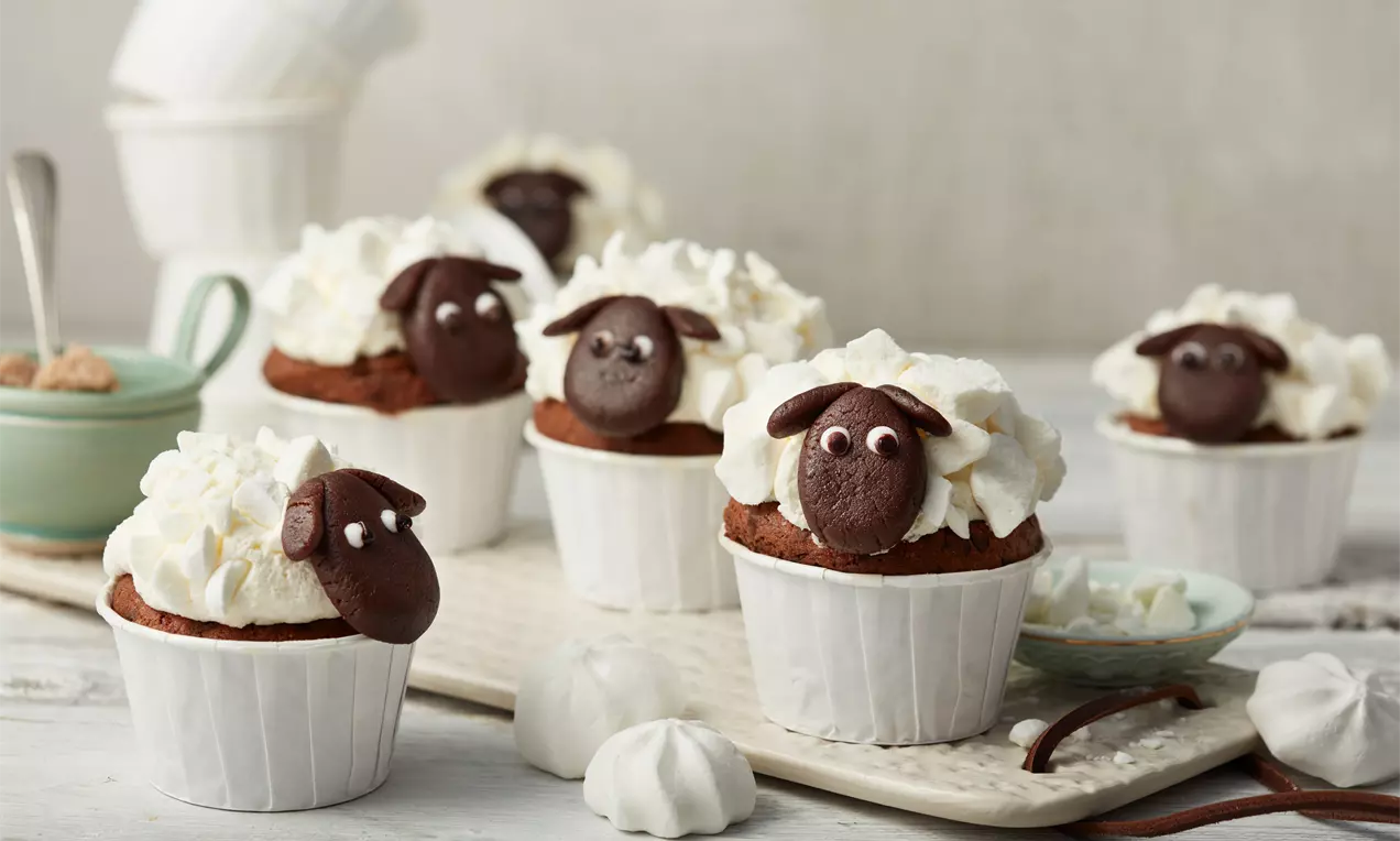 Schäfchen-Cupcakes aus dunklem Teig mit weißer Sahne dekoriert, Schokoladengesicht und Augen, angerichtet auf einem rechteckigen Tablett