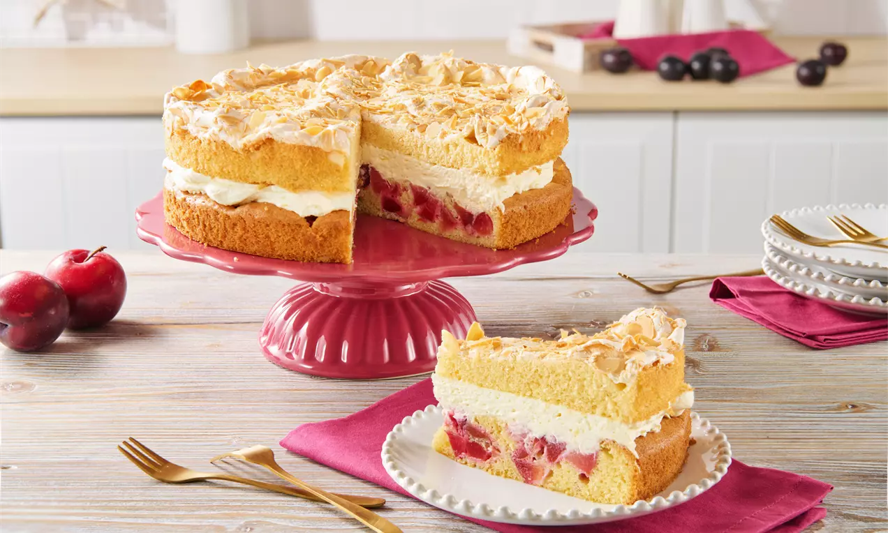Pflaumen-Baiser-Torte mit goldgelbem Boden, weißer Creme, fruchtiger Pflaumenfüllung und luftiger Baiserschicht auf roter Tortenplatte