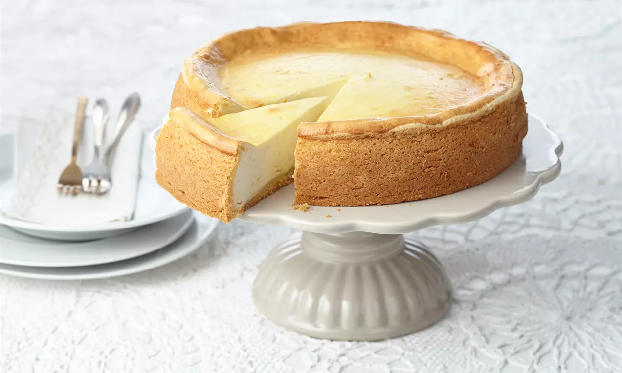 Goldbrauner Käsekuchen mit glatter Füllung und leicht gebräunter Oberfläche, auf einer stilvollen Kuchenplatte mit Teller und Besteck im Hintergrund