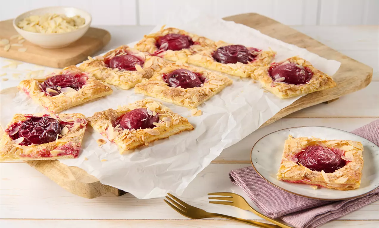 Goldbraune rechteckige Blätterteigteilchen mit saftigen Pflaumenhälften und Mandelblättchen auf Backpapier, daneben ein Teller mit Serviette