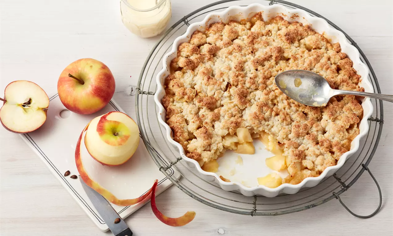 Goldbrauner Obst-Crumble in weißer Backform mit saftigen Apfelstücken, flankiert von frischen Äpfeln und einem Glas heller Sauce