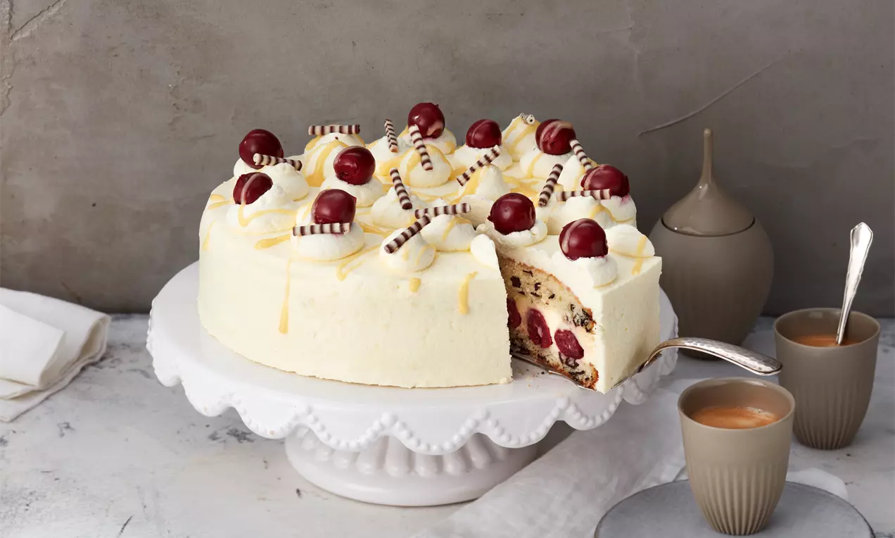 Eierlikörtorte mit hellgelber Creme, Kirsch-Dekoration, Schokoröllchen und Anschnitt auf weißer Tortenplatte mit Kaffee und Keramikvase