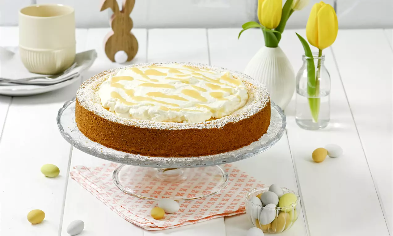 Eierlikör-Käsekuchen mit goldbrauner Kruste, weißer Sahneschicht und Eierlikörmuster auf einer Glasplatte mit Frühlingsdeko angerichtet