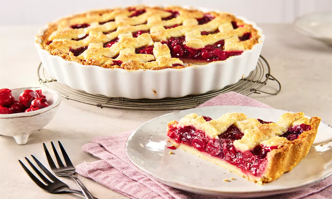 Ein goldbrauner Kirschkuchen mit Gitterkruste und roter Füllung in weißer runder Form, daneben ein Stück auf Teller mit rosa Stoffserviette