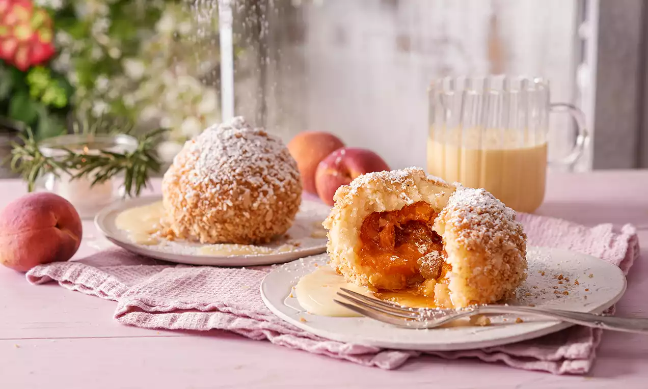 Zwei goldbraune Aprikosenknödel mit knuspriger Bröselhülle, einer geöffnet und gefüllt mit orangefarbener Aprikose, auf hellem Teller bestäubt mit Puderzucker und umgeben von rosa Dekoelementen und Aprikosen