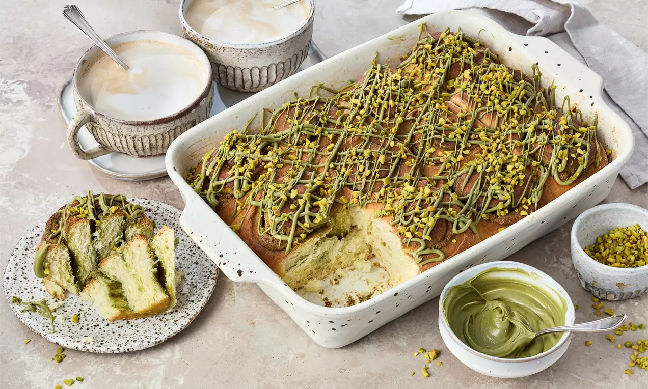 Goldbraune Pistazienschnecken in einer weißen Keramikform, mit grünem Pistazienmus bestrichen und gehackten Pistazien bestreut, daneben Tassen mit Milchschaum