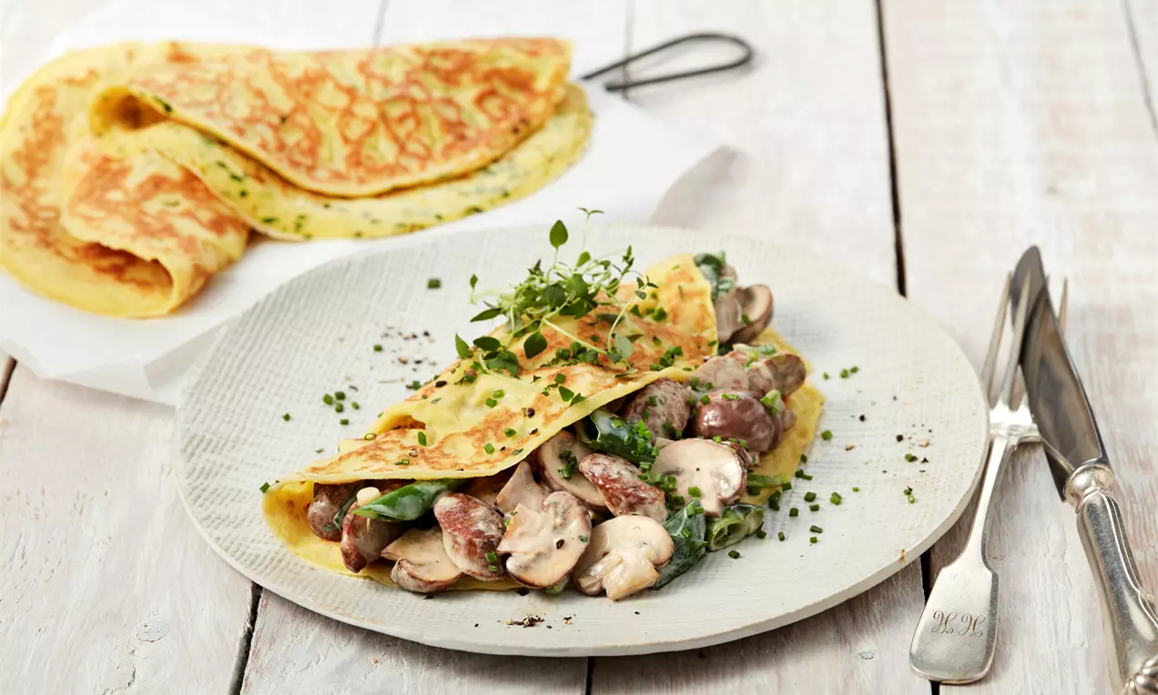 Dünner goldgelber Pfannkuchen mit grünen Kräutern gefüllt mit braunen Champignons und Würstchen, angerichtet auf hellem Teller mit frischen Kräutern