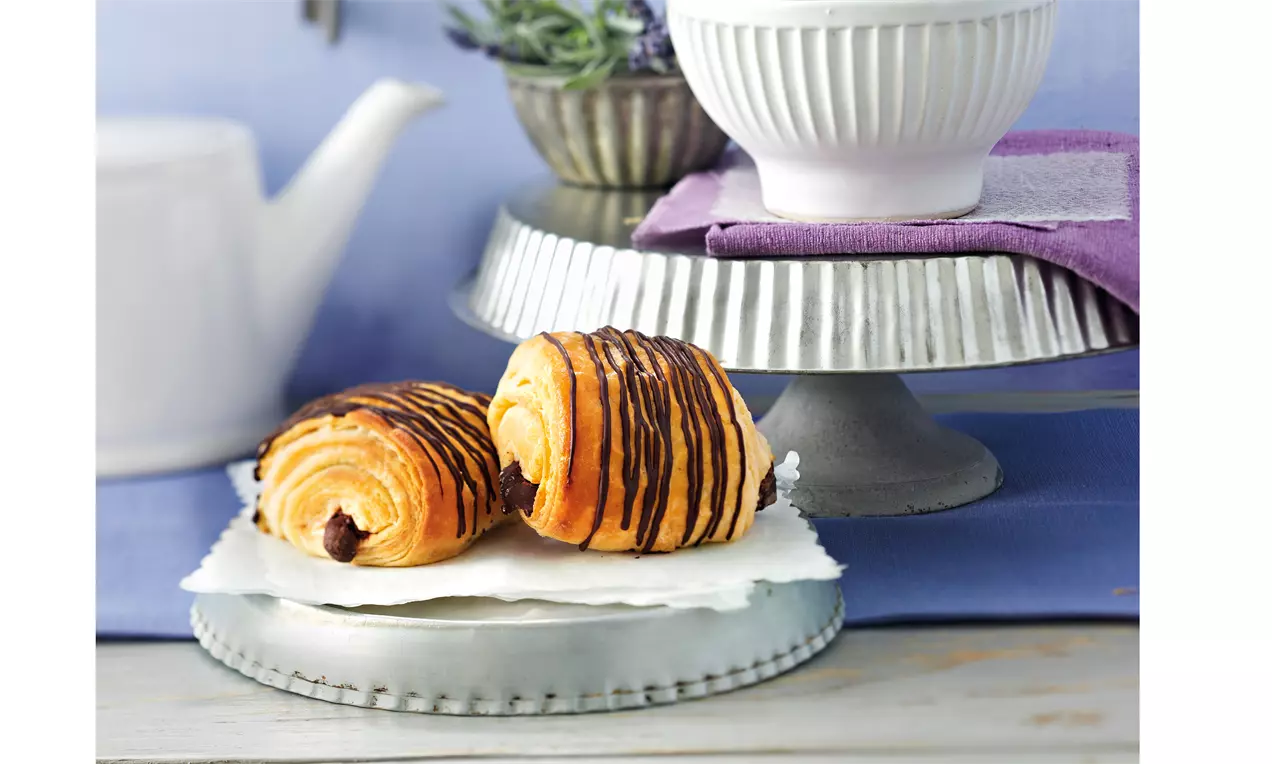 Goldbraune Pains au chocolat mit geschwungenen Schichten und dunkler Schokoladenfüllung, dekoriert mit feinen Schokoladenstreifen auf weißer Serviette