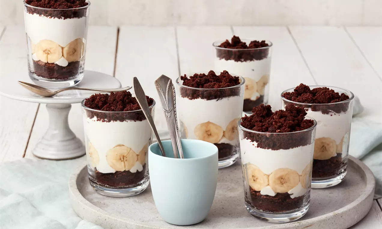 Schichtdesserts im Glas mit dunklem Schokoboden, Bananenscheiben, heller Creme und Schokokrümel-Topping auf grauem Tablett arrangiert